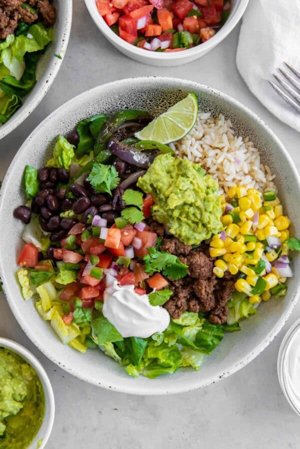 A white bowl filled with rice, cooked ground beef, black beans, corn salsa, diced tomatoes, guacamole and sour cream to make a beef burrito bowl.