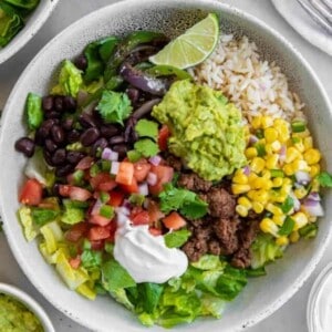 A white bowl filled with rice, cooked ground beef, black beans, corn salsa, diced tomatoes, guacamole and sour cream to make a beef burrito bowl.