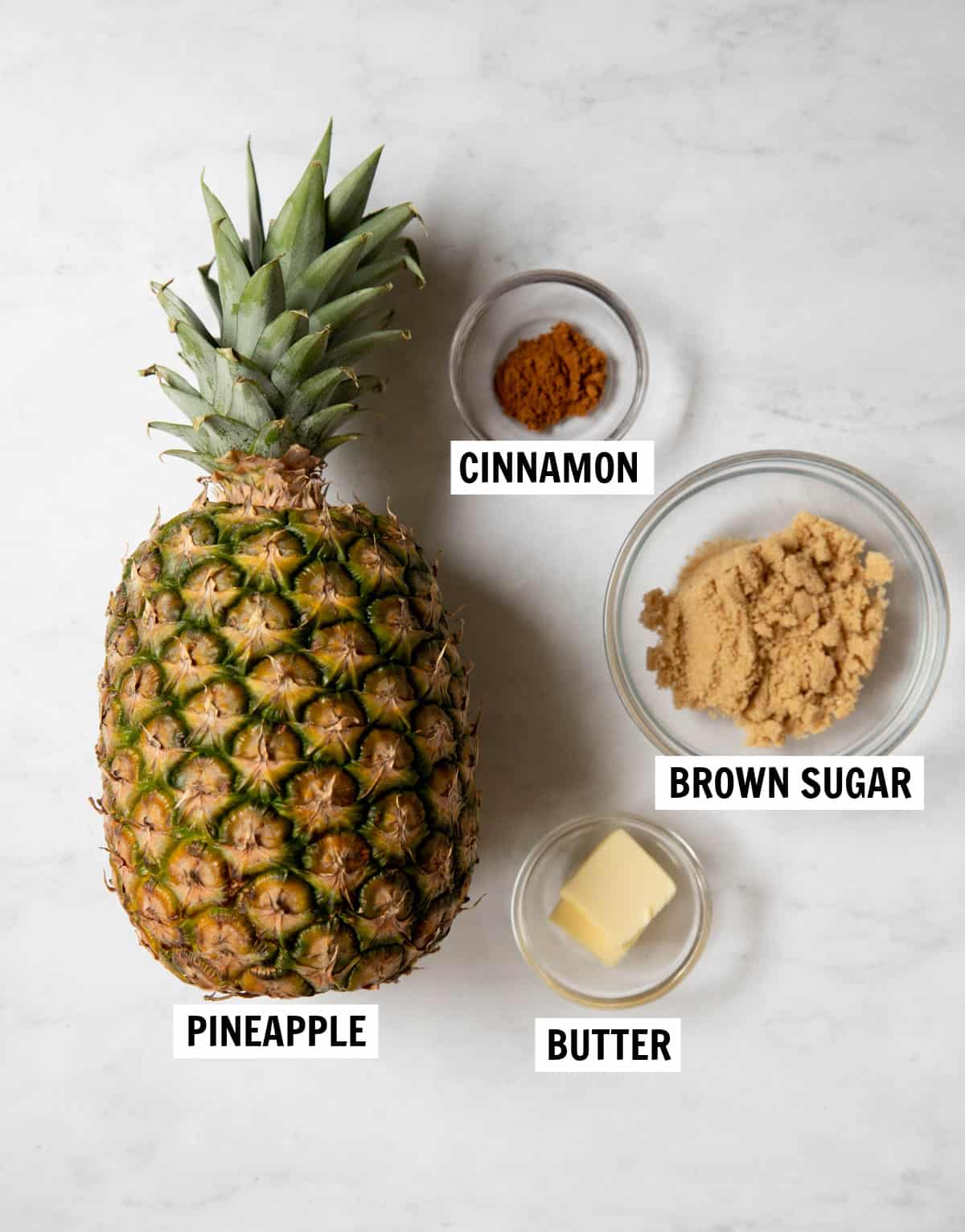 All of the ingredients for fried pineapple on a white countertop including a whole pineapple, butter, cinnamon and brown sugar.