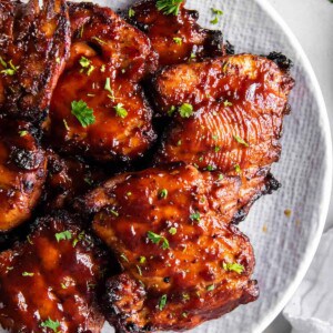 A white plate stacked with bbq boneless chicken thighs ready for serving. They are garnished with fresh chopped parsley.