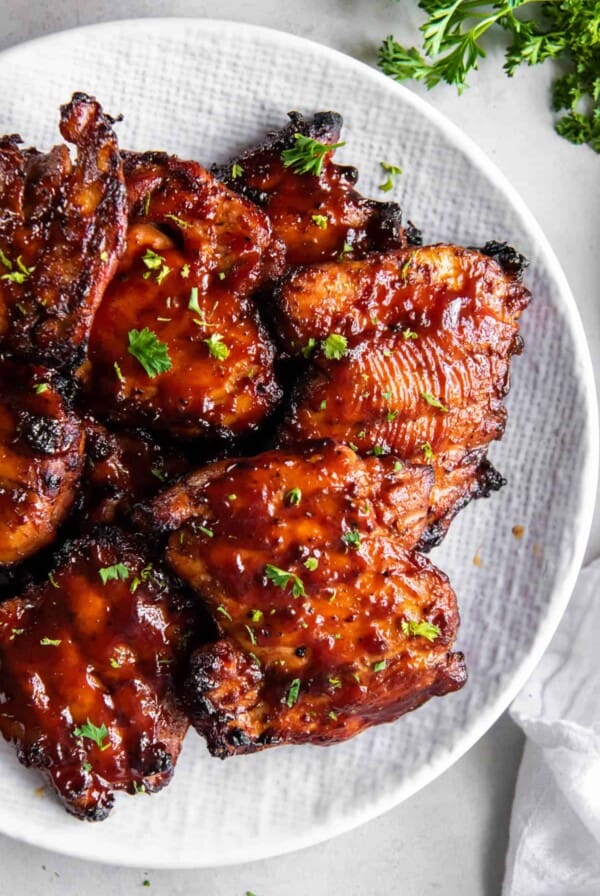 A white plate stacked with bbq boneless chicken thighs ready for serving. They are garnished with fresh chopped parsley.