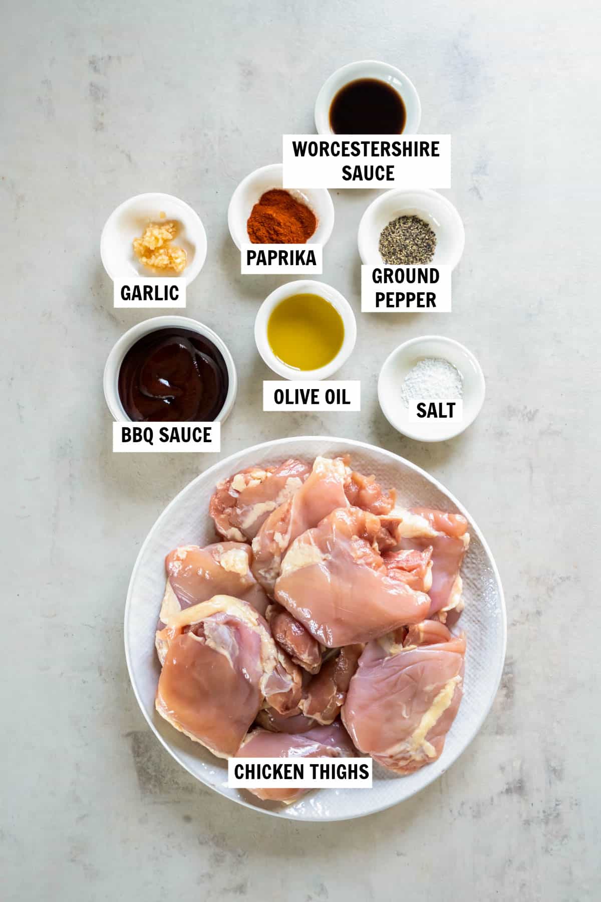 All of the ingredients for bbq boneless chicken thighs in bowls on a white countertop including chicken thighs, olive oil, BBQ sauce, garlic, Worcestershire sauce, paprika, kosher salt and black pepper.