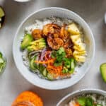 Shrimp and rice bowl in a white bowl including the cooked rice layered in the bowl with the other ingredients like the cooked shrimp, avocado, pineapple, pickled carrots and sliced cucumbers.