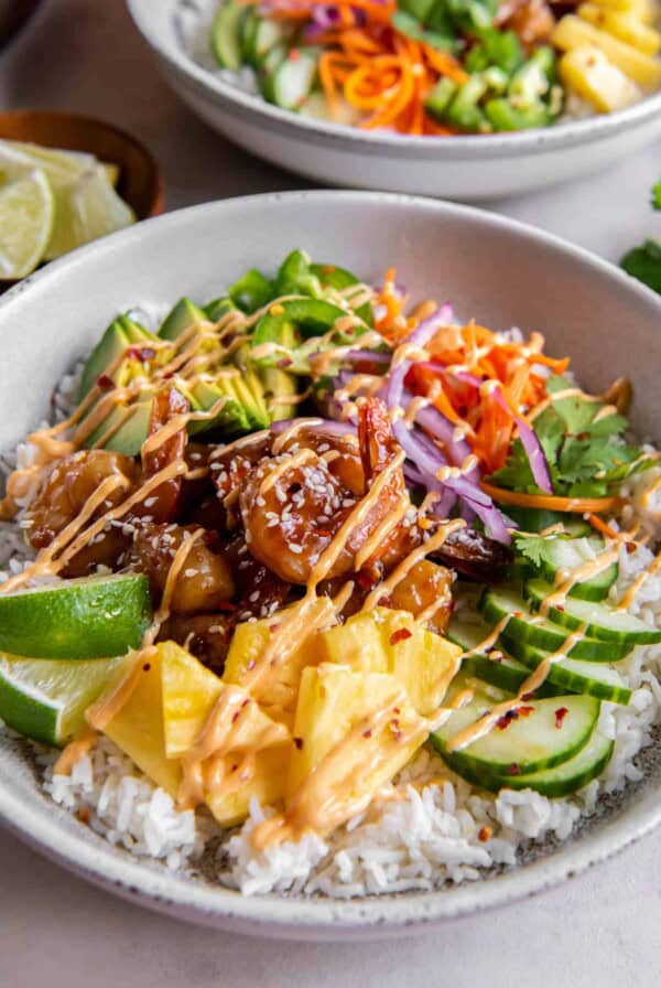 A white bowl with rice and piled high with pineapple, cucumber, carrots, shrimp, red onion and avocado. Everything is drizzled with ariracha mayo.