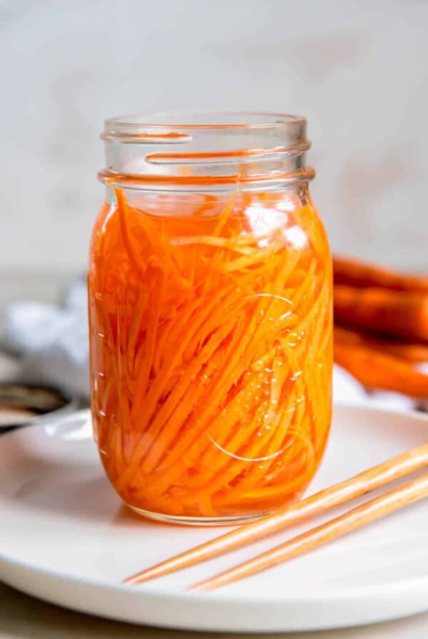 An open jar of quick picked carrots sitting on a white plate on a countertop.