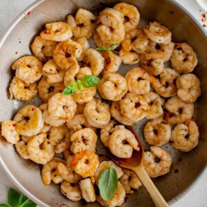 Pan seared shrimp in a steel skillet after cooking. The cooked shrimp is garnished with fresh basil leaves and a wooden spoon sits in the pan to serve the shrimp.