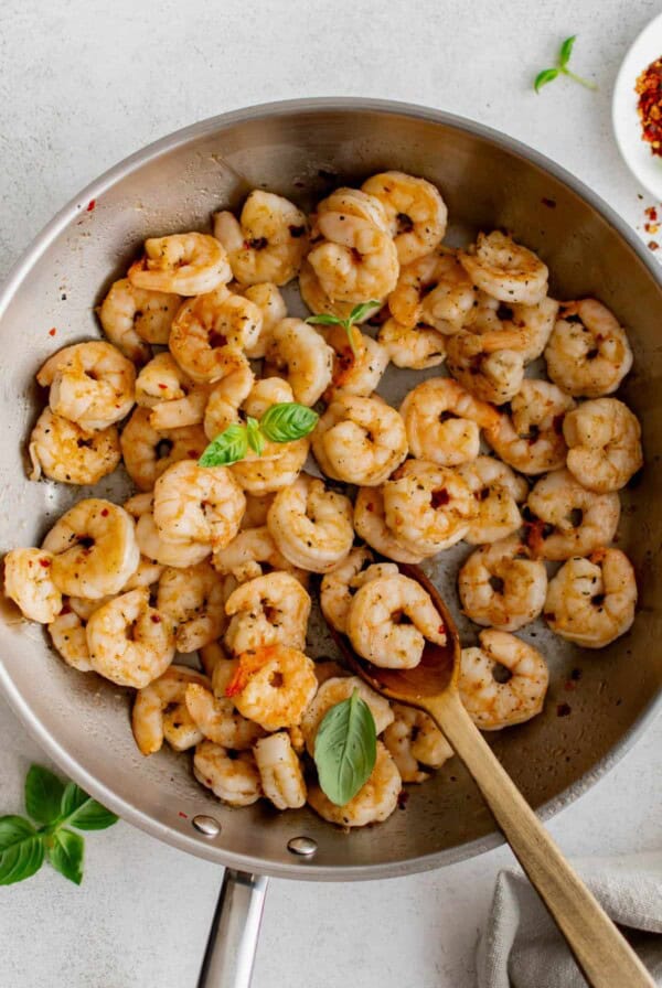 Pan seared shrimp in a steel skillet after cooking. The cooked shrimp is garnished with fresh basil leaves and a wooden spoon sits in the pan to serve the shrimp.