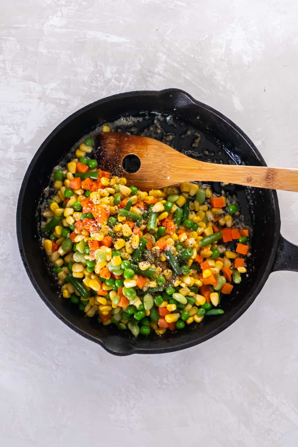 A spoon sits in a pan with the mixed vegetables and salt and pepper is sprinkled on top for mixing together before serving.