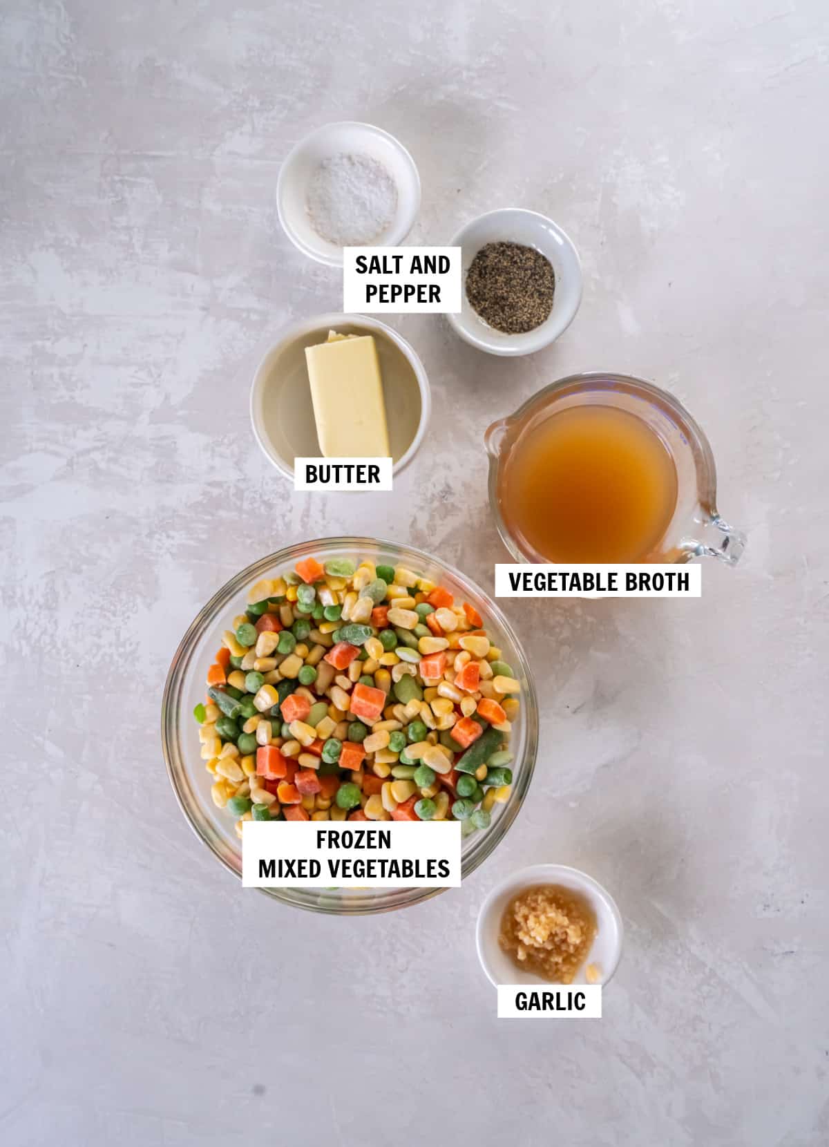 All of the ingredients for buttered mixed vegetables in bowls on a white countertop including frozen mixed vegetables, vegetable broth, garlic, butter, salt and pepper.