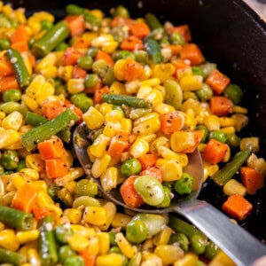 A spoon sits in a cast iron pan filled with buttered mixed vegetables ready for serving.