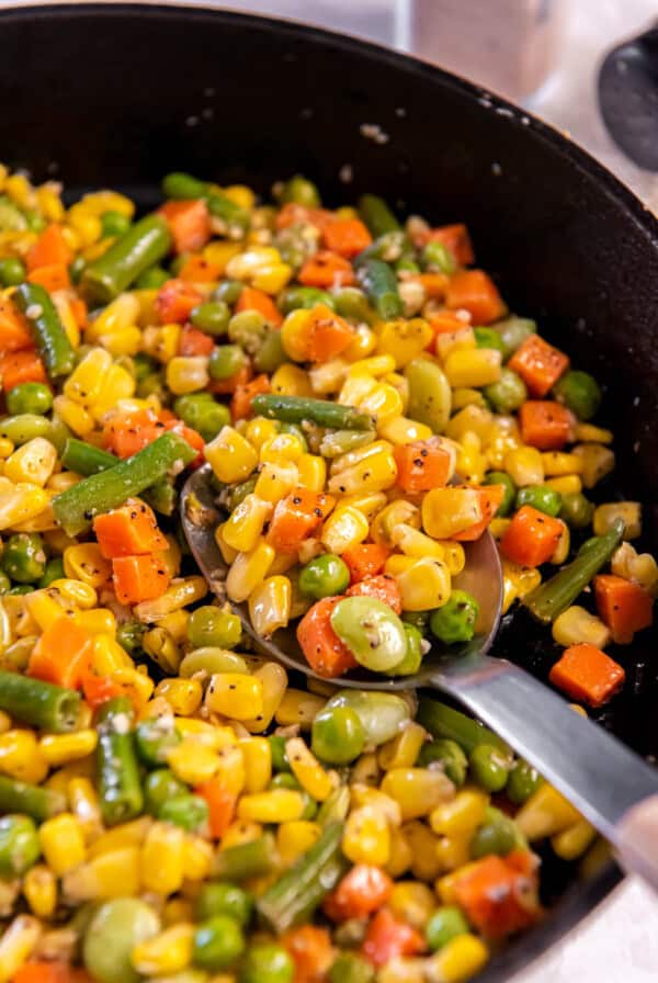 A spoon sits in a cast iron pan filled with buttered mixed vegetables ready for serving.