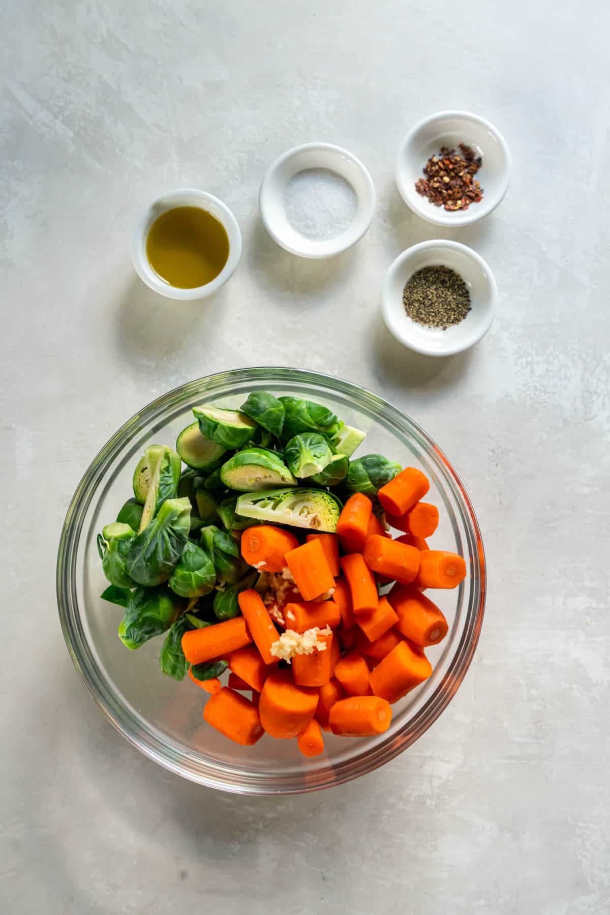 Ingredients for roasted vegetables sitting in bowls on a countertop including carrots, brussels sprouts, salt, pepper, olive oil and spices.