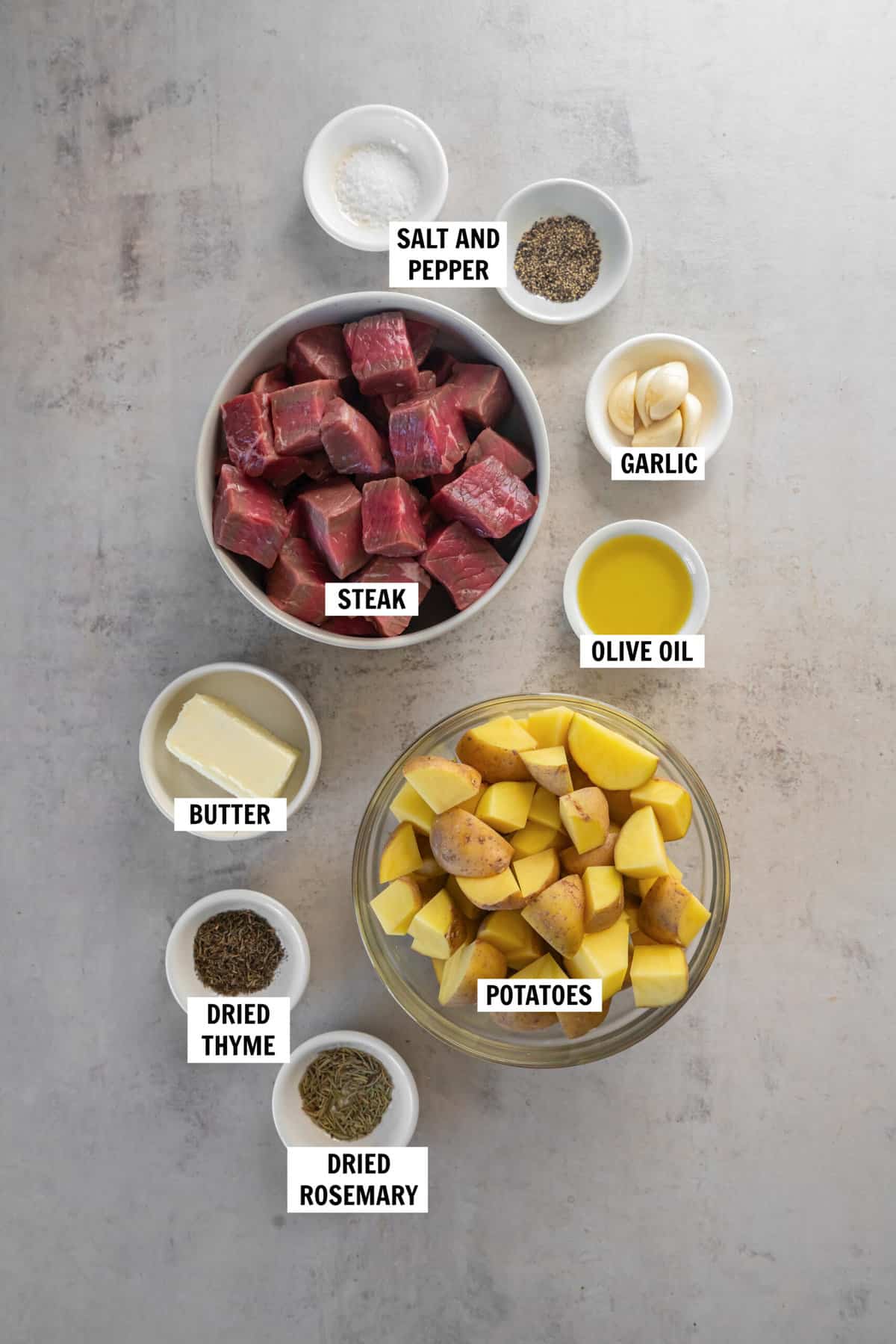 All of the ingredients for steak bites and potatoes in bowls on a light countertop including steak, potatoes, olive oil, garlic, salt and pepper, butter, dried rosemary and dried thyme.