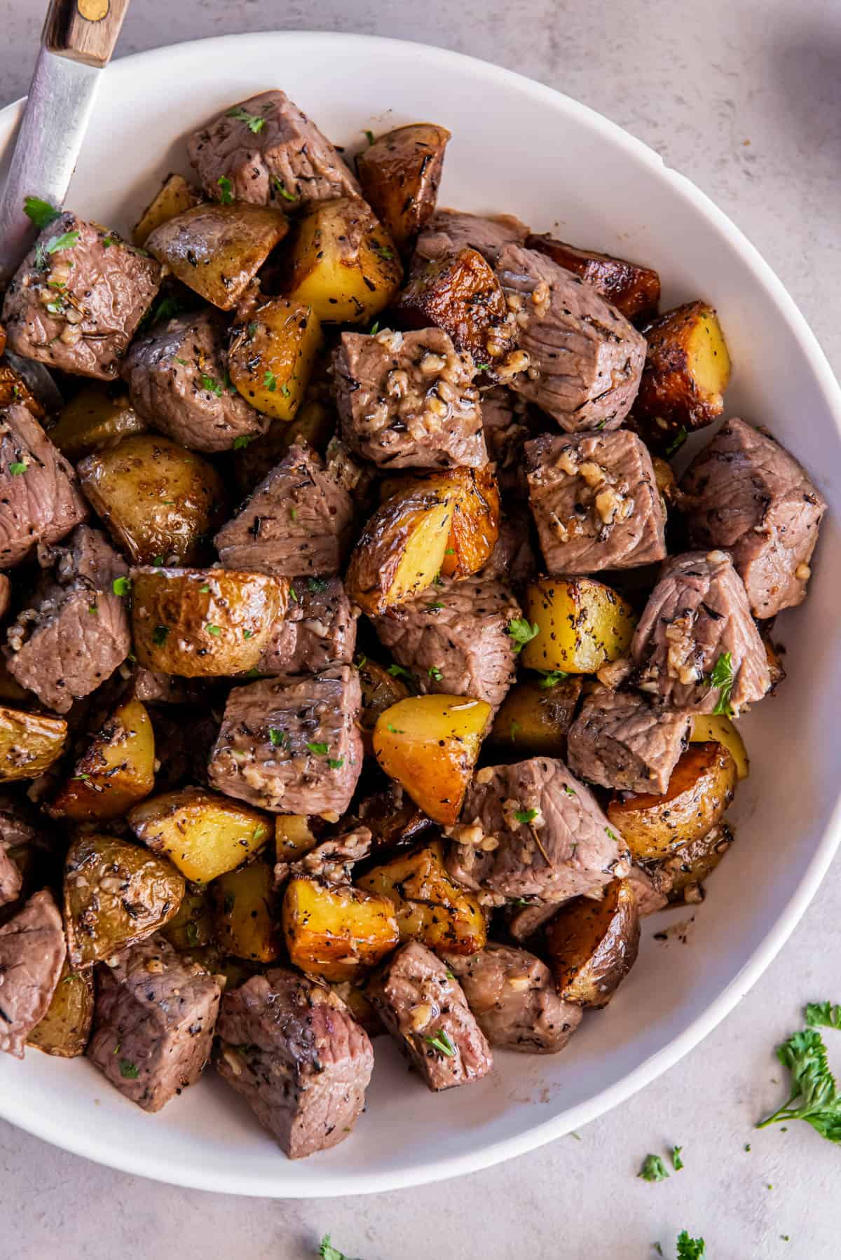 Garlic steak bites and potatoes in a white serving bowl for serving. Freshly chopped parsley is sprinkled on top. 