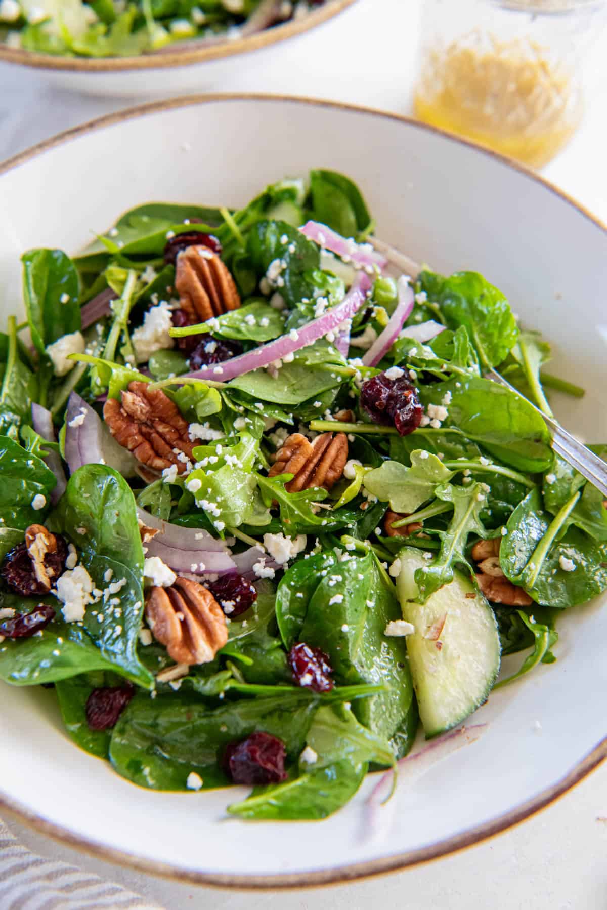 A light and refreshing spinach and arugula salad sits in a white bowl for serving. The spinach and arugula is topped with cucumber, dried cranberries, red onion, pecans,. feta cheese and dressing.