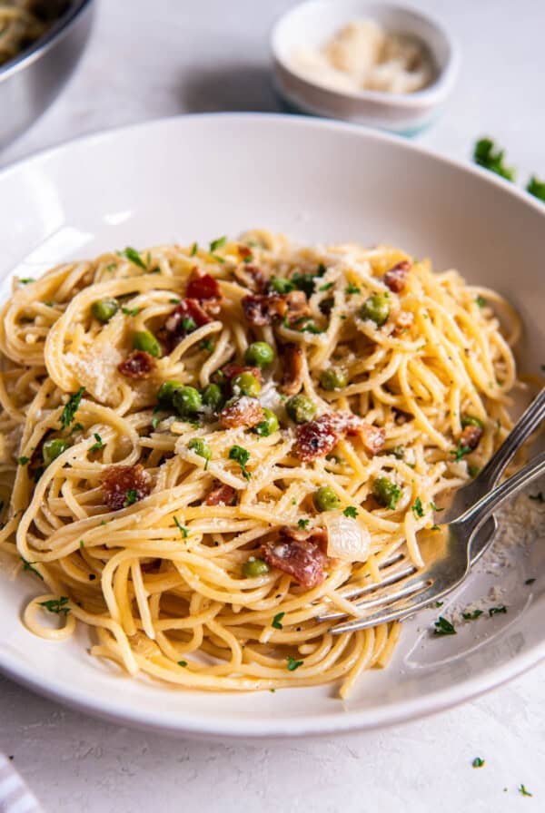 A white bowl filled with pasta with peas and bacon. A spoon rests in the bowl for serving.