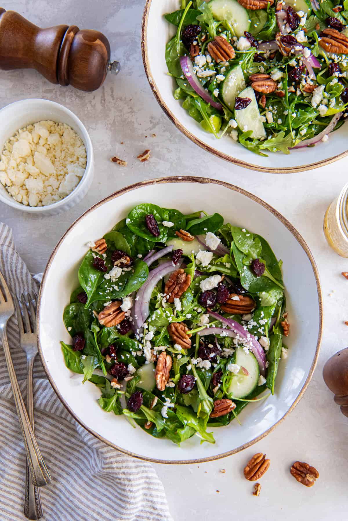 Two bowls filled with spinach arugula salad on a white tabletop for serving. A bowl of crumbled feta and cracked black pepper sits to the side of the salad bowls. 