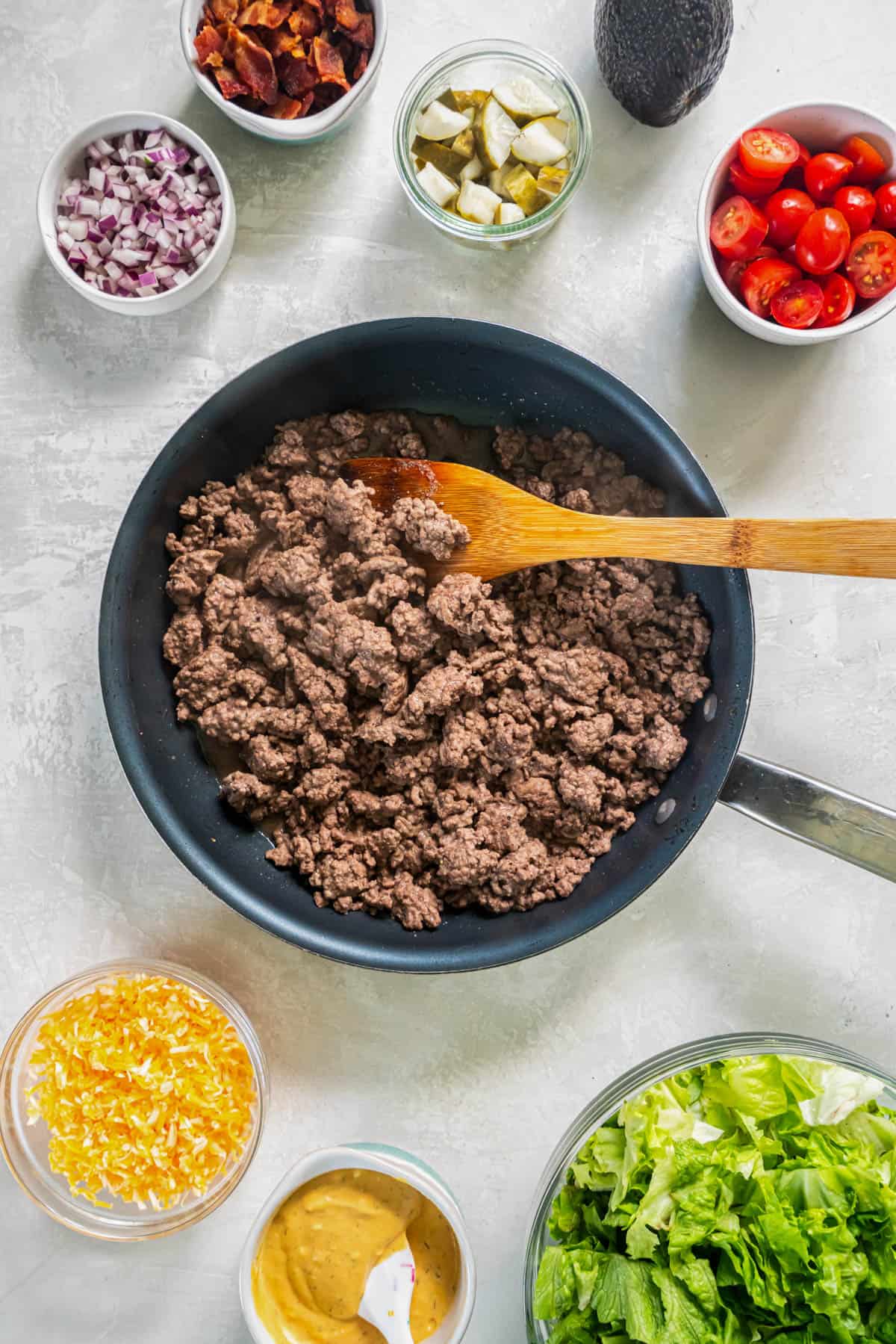 Cooked beef in a skillet with a wooden spoon sitting on a countertop with all of the other burger bowl toppings sitting in bowls on a countertop.