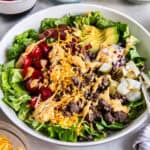 A white bowl filled with lettuce, cooked ground beef, tomatoes, avocado, pickles, cooked bacon, red onion and shredded cheese.