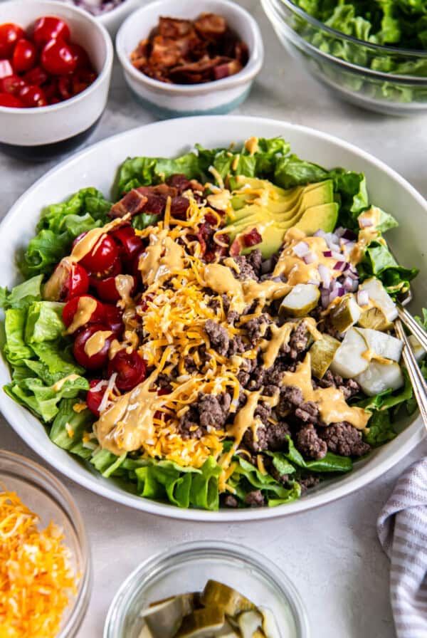 A white bowl filled with lettuce, cooked ground beef, tomatoes, avocado, pickles, cooked bacon, red onion and shredded cheese.