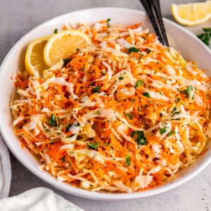 Cabbage and carrot salad in a white bowl on a countertop with two serving spoons.