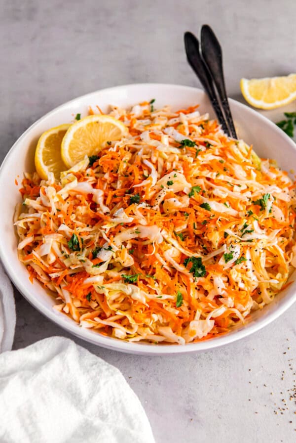 Cabbage and carrot salad in a white bowl on a countertop with two serving spoons.