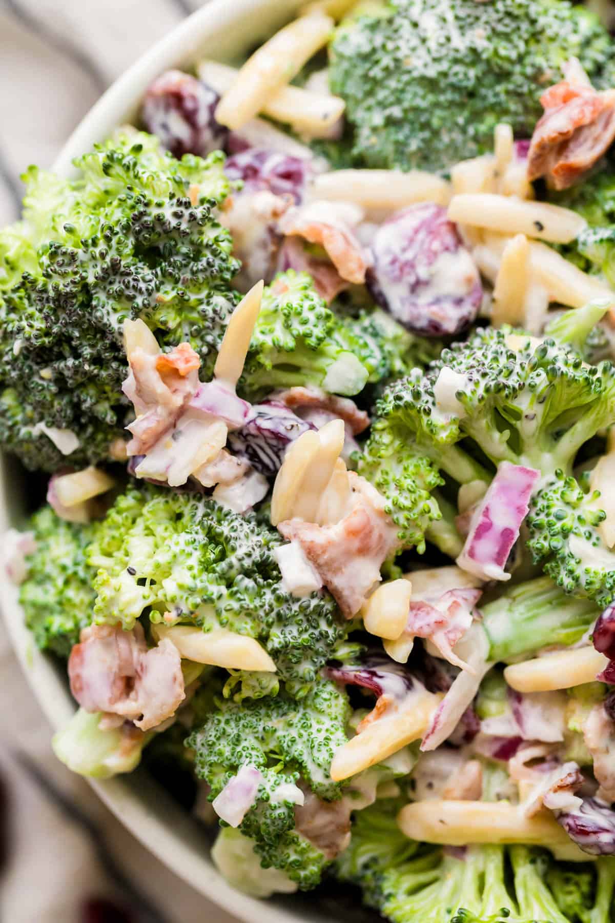 A close up photo of broccoli crunch salad in a bowl ready for serving including broccoli florets, diced red onion, slivered almonds and dried cranberries. 