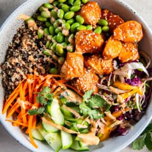 A Salmon Quinoa Bowl is loaded with sweet chili glazed salmon, mango slaw, shredded carrots, cucumber, edamame, fresh cilantro and spicy mayo.