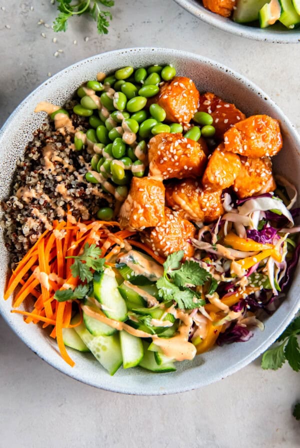 A Salmon Quinoa Bowl is loaded with sweet chili glazed salmon, mango slaw, shredded carrots, cucumber, edamame, fresh cilantro and spicy mayo.