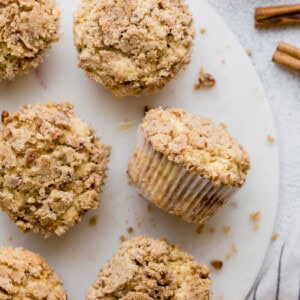 Six coffee cake muffins sit on a white serving platter with crumbs on the side.