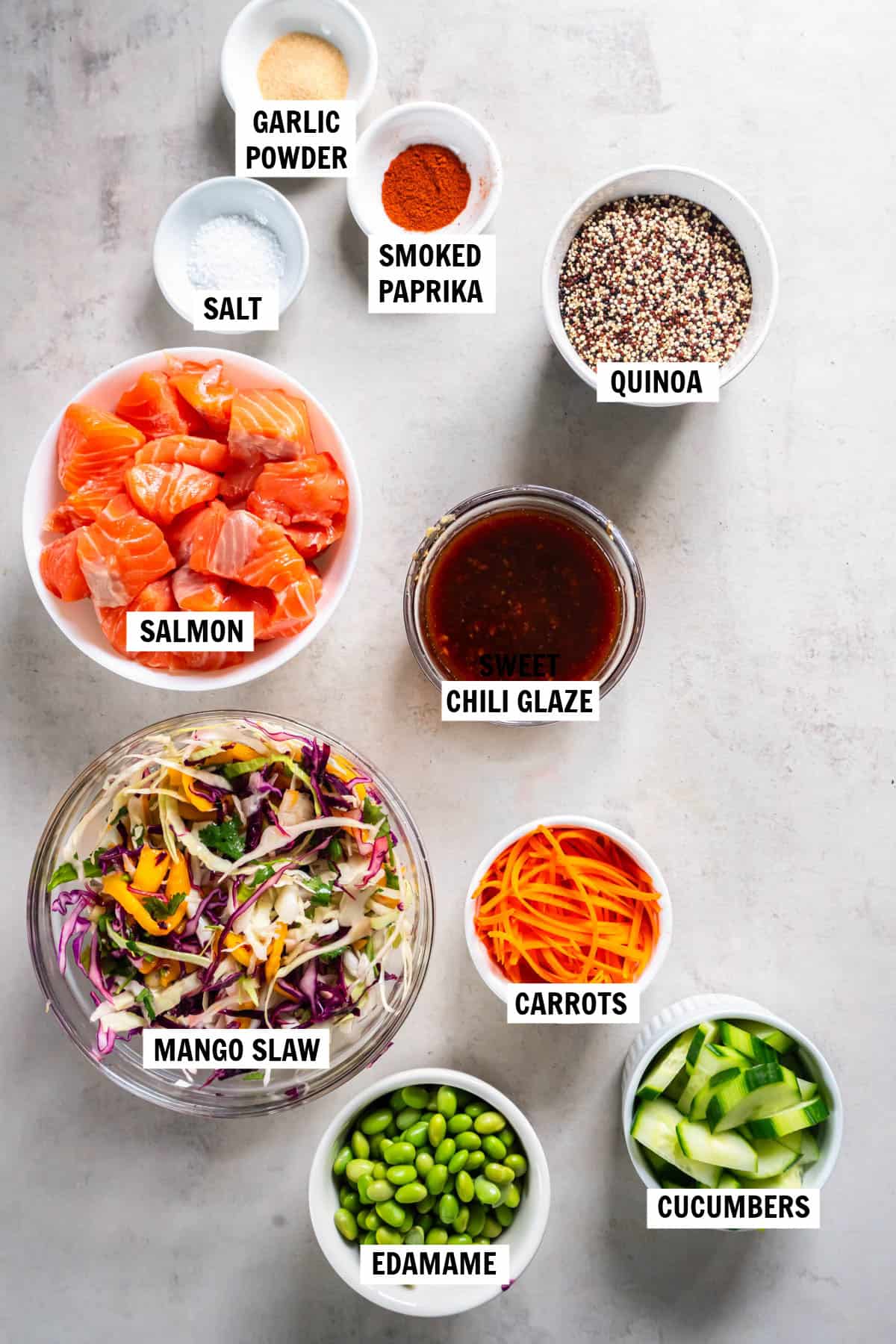 The ingredients for salmon quinoa bowl in bowls on a countertop including salmon, mango slaw, edamame, carrots, cucumbers, quinoa, sweet chili sauce, garlic powder, smoked paprika, and salt. 