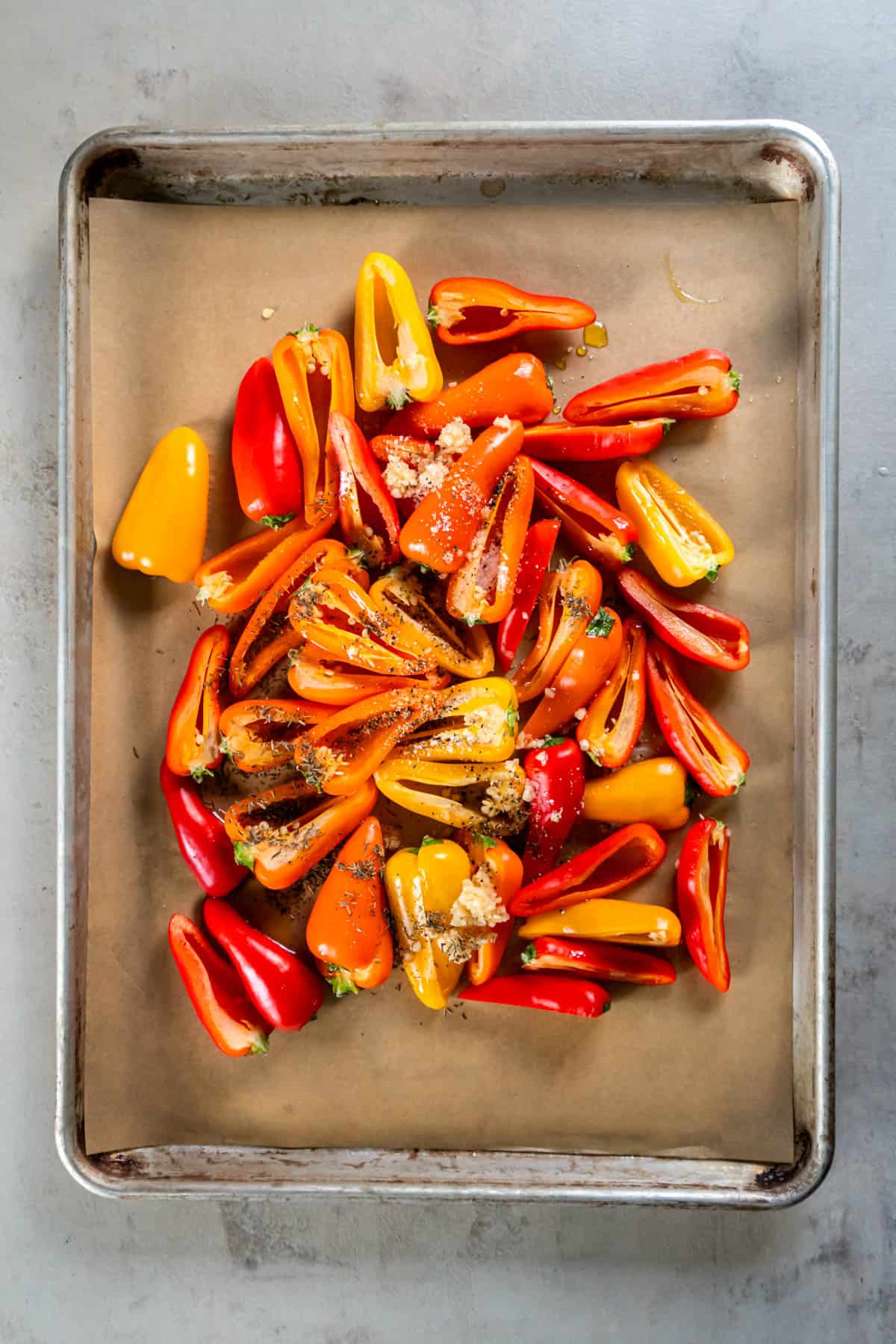 Mini peppers split in half and scattered on a baking sheet lined with parchment paper. Olive oil, garlic, dried thyme, salt and peppers is sprinkled on top before roasting in the oven. 