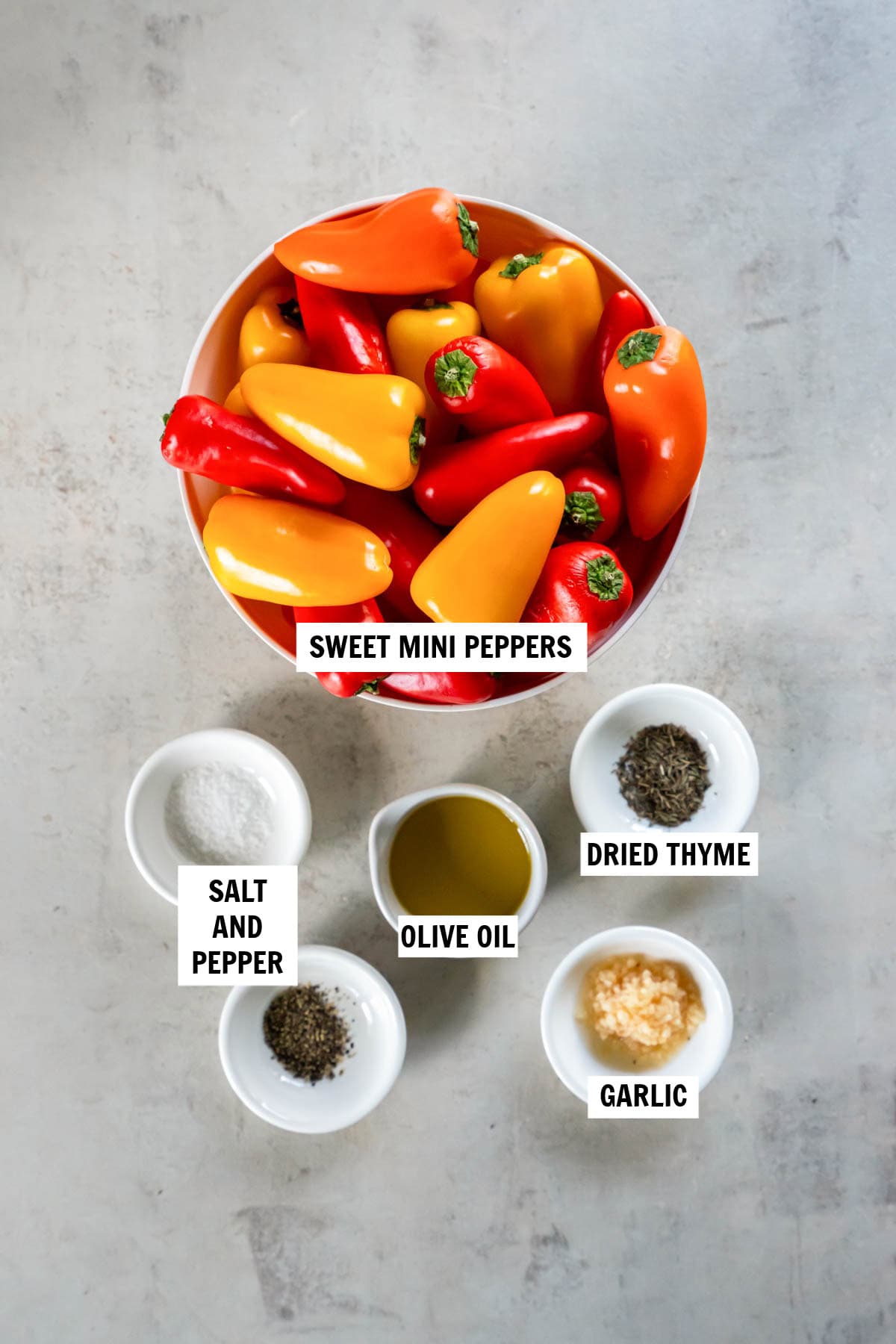 All of the ingredients for roasted mini peppers in bowls on a white countertop including mini sweet peppers, olive oil, dried thyme, garlic, salt and pepper.