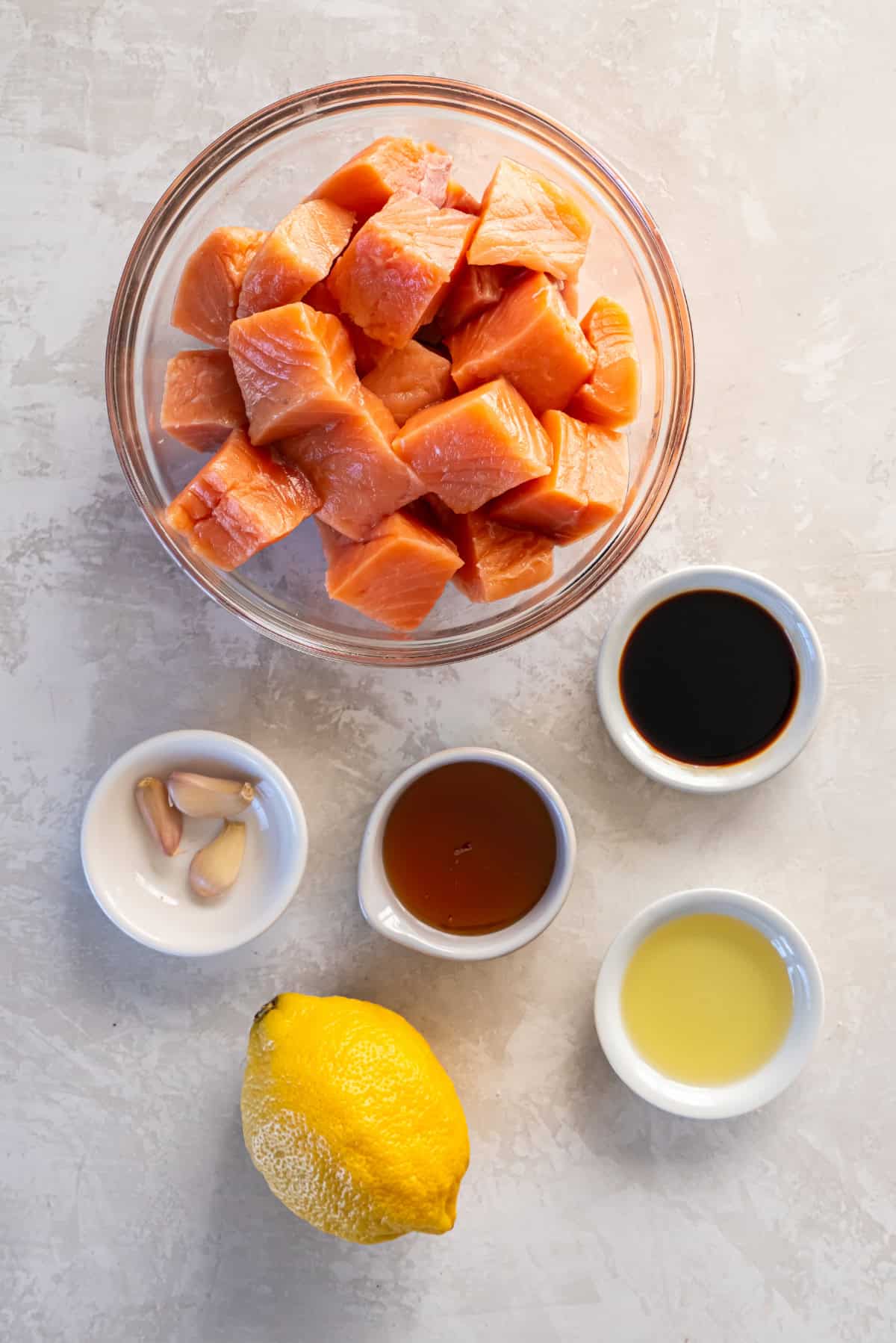 The ingredients for honey garlic salmon bites in bowls. There is diced salmon, soy sauce, honey, garlic cloves, lemon juice, and a lemon.