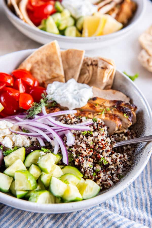 A Mediterranean Chicken Bowl with cucumbers, tomatoes, red onion, quinoa, pita wedges, tzatziki sauce, and cooked chicken.