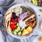 A Mediterranean Chicken Bowl with cucumbers, tomatoes, red onion, quinoa, pita wedges, tzatziki sauce, and cooked chicken.