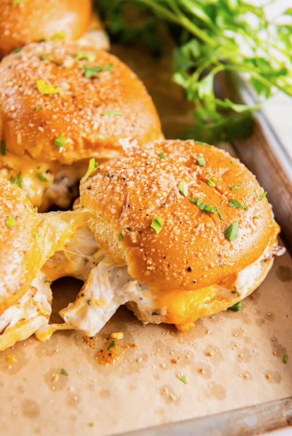 A Hawaiian roll sandwich with chicken and melted cheese in it. It is sprinkled with parmesan cheese. It is on a tray with parchment paper.