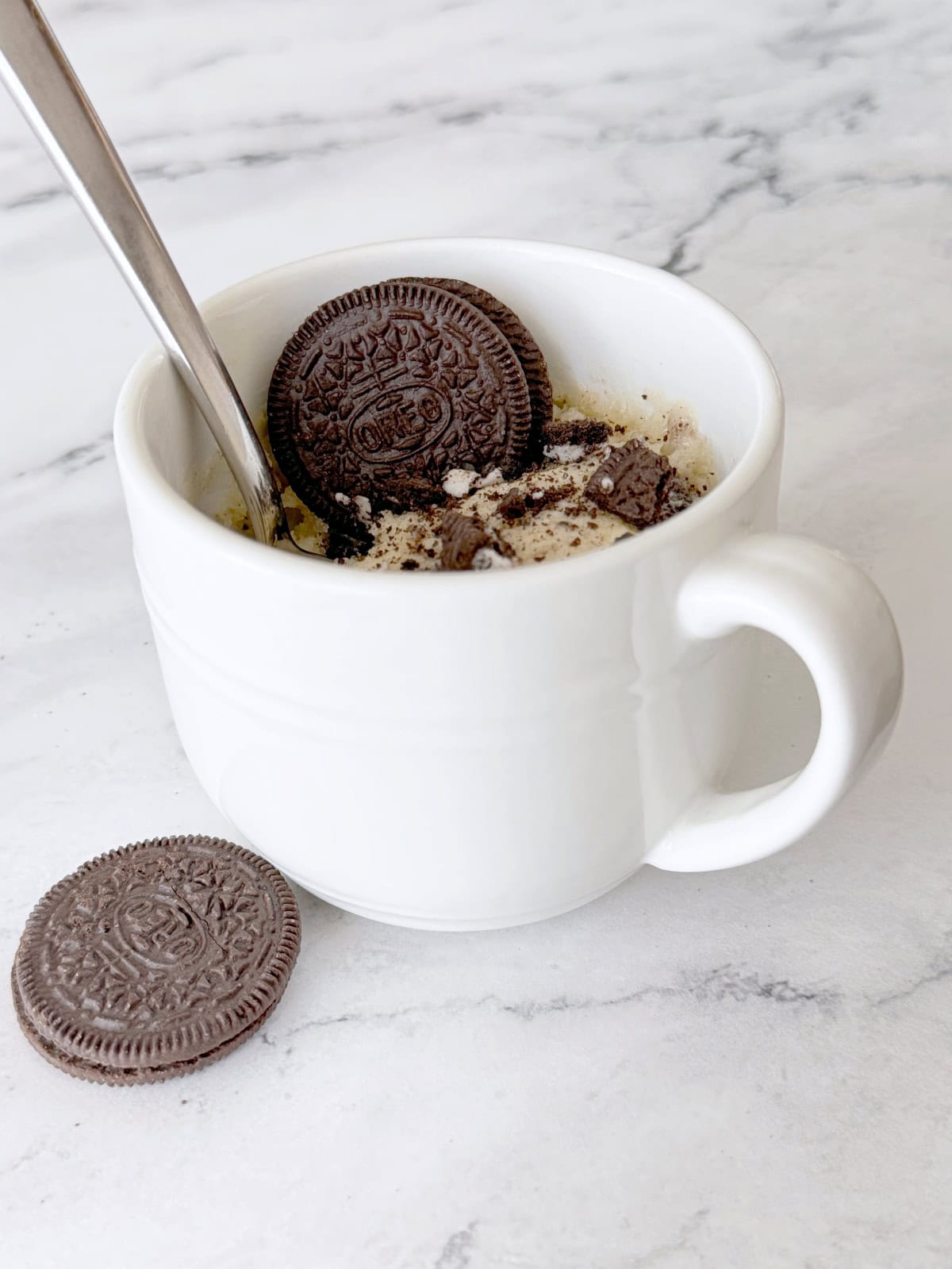 a white mug with cake inside it. A spoon is sitting in the mug and an Oreos resting in the mug and on the counter.